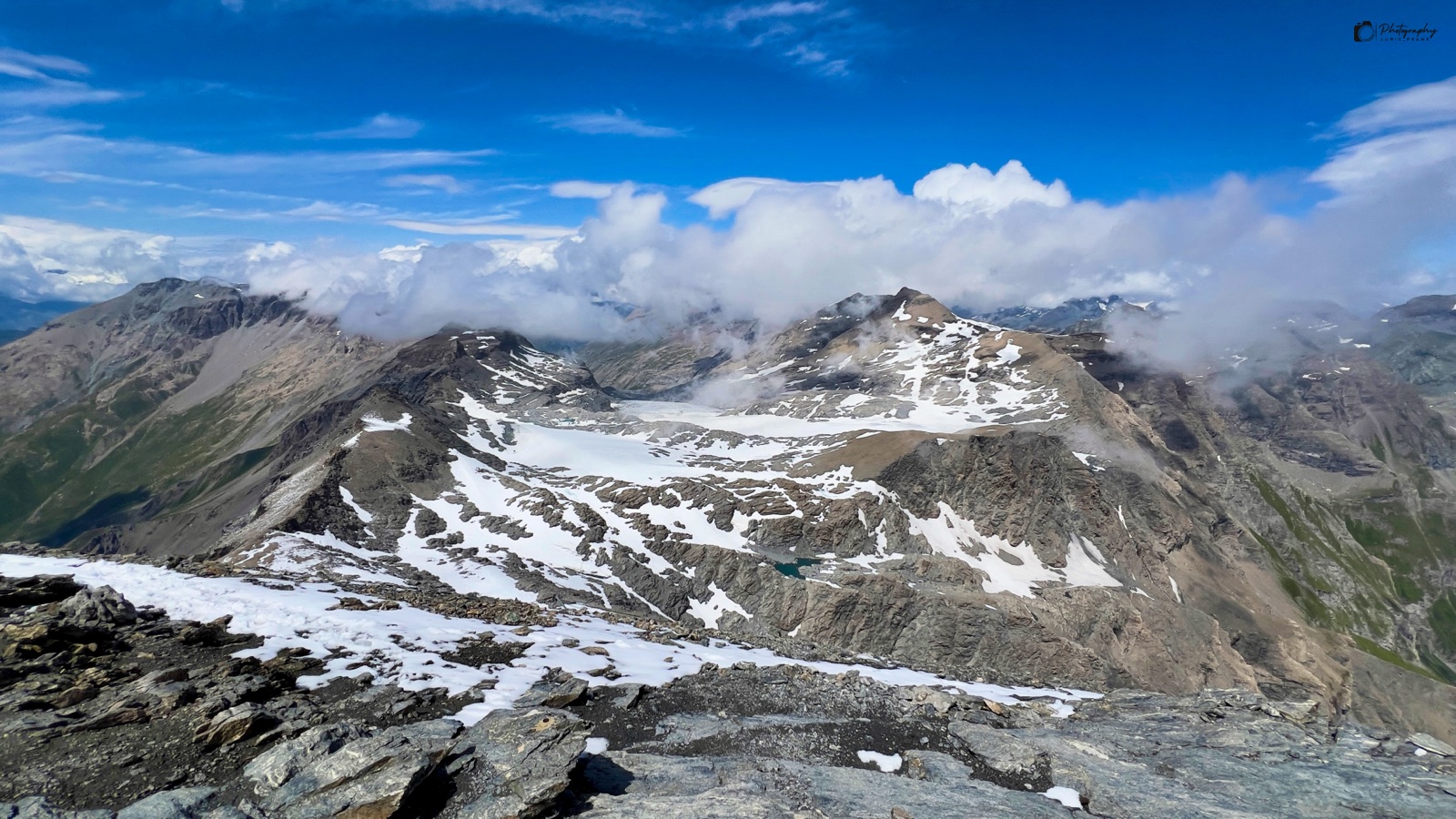 Montagne con chiazze di neve sotto un cielo azzurro, con nuvole che avvolgono le cime