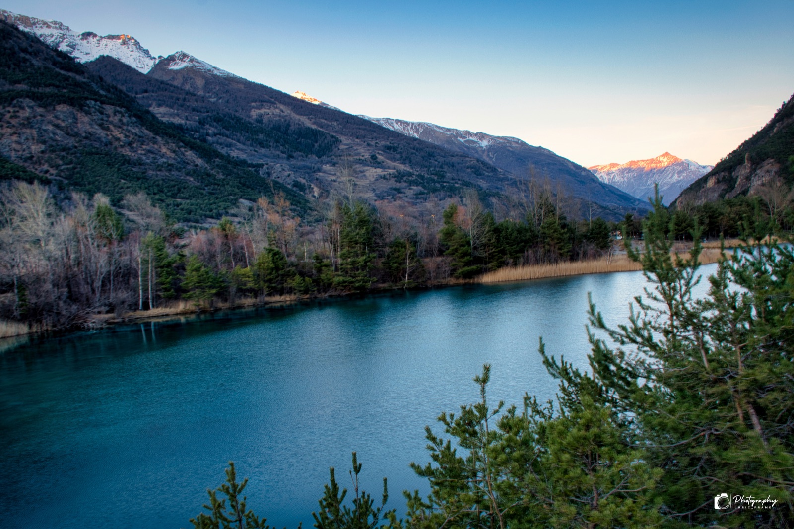 Lago dalle acque blu ai piedi di montagne innevate, con alberi sulla riva nella luce della sera