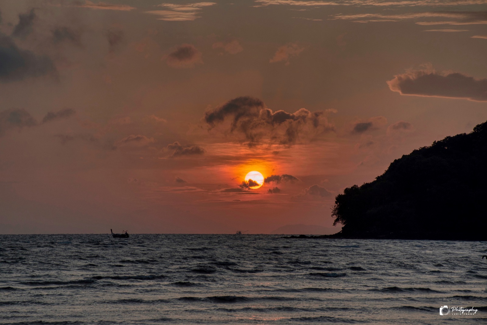 Sole che tramonta tra le nuvole sopra il mare, con una piccola barca all'orizzonte e un promontorio scuro sulla destra