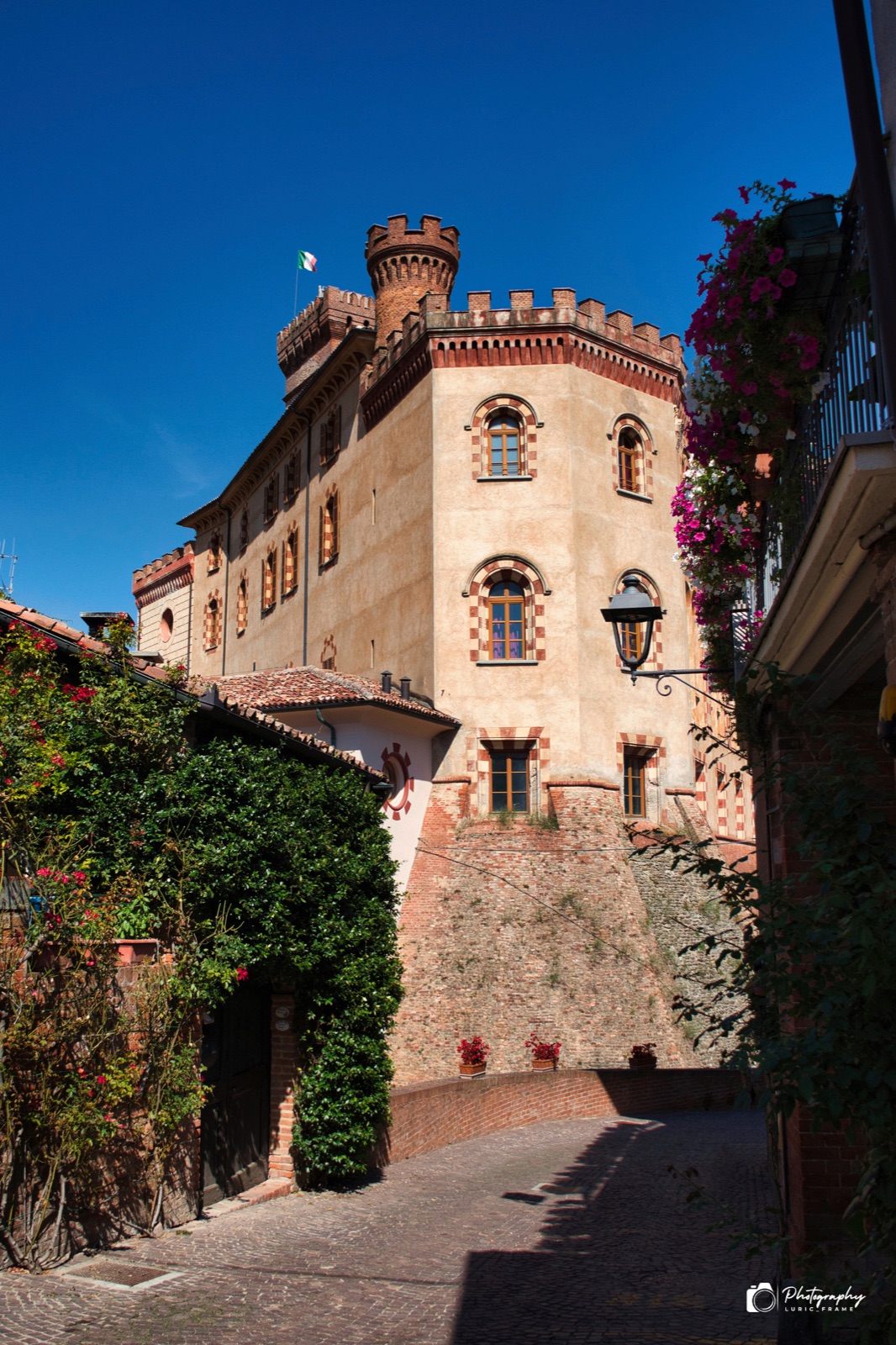 Castello in mattoni con torri merlate visto da una stretta via in salita, tra piante rampicanti e fiori